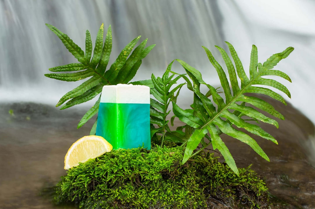 Photographie d'un savon mis en scène au pied d'une cascade avec de la mousse, des fougères et une tranche de citron
