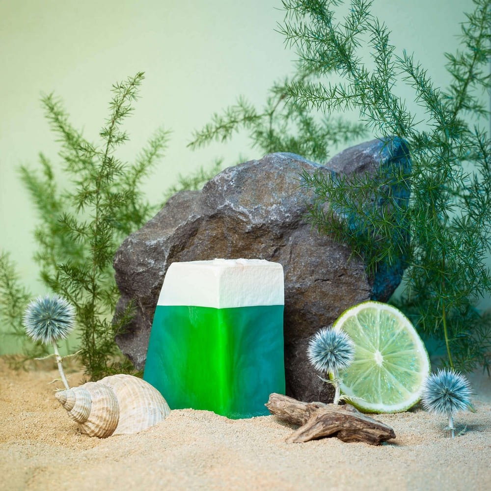 Photographie d'un savon mis en scène dans un décor de plage, avec du sable, coquillage, citron, fleurs et plantes